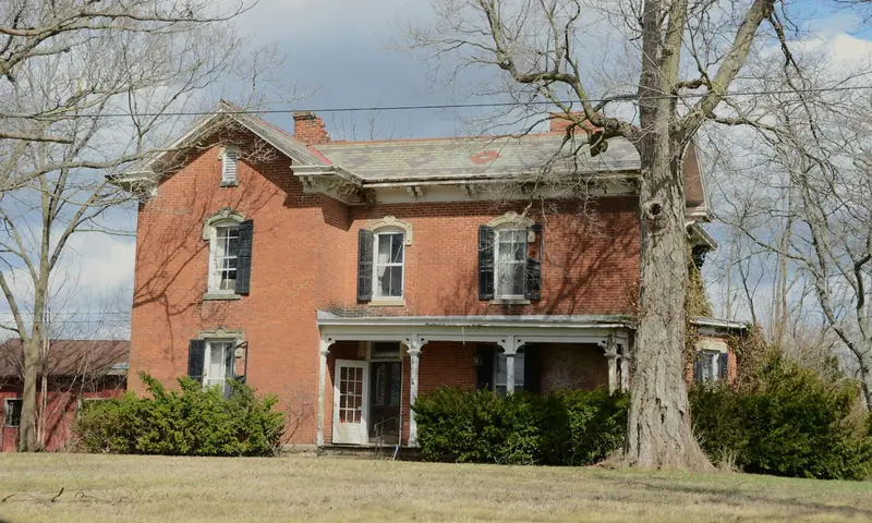Older red brick farmhouse with shutters and a side porch under tall trees
