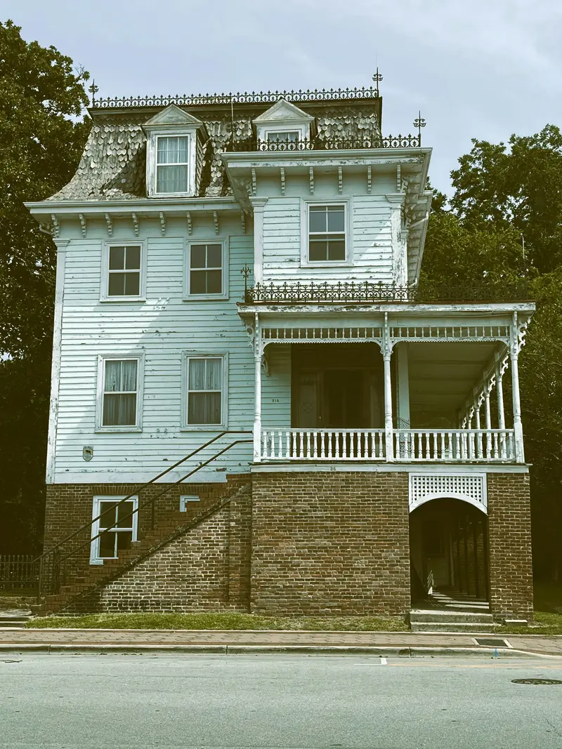 Pale blue three-story Victorian house with an ornate white porch over a brick base